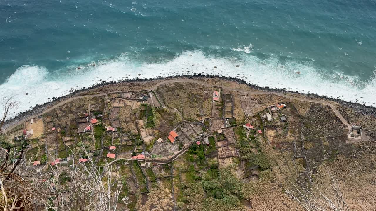 Aerial view of a coastal village with scattered houses, farmland, and ocean waves crashing onto the rugged shoreline. Madeira Island, Portugal.