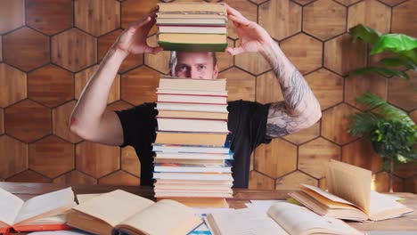 young caucasian man have fun hiding eyes behind mountain of books sits at table