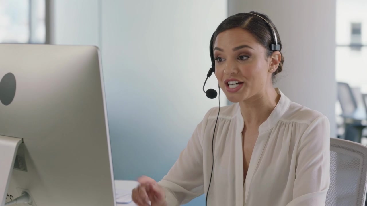 Rappresentante del servizio clienti con camicetta bianca che parla in un auricolare in ufficio per assistere un cliente durante una chiamata.