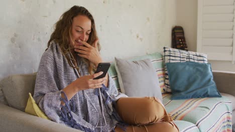 Mujer-Caucásica-Sonriente-Con-Manta,-Sentada-En-Un-Sofá-Usando-Un-Teléfono-Inteligente-En-La-Sala-De-Estar-De-La-Cabaña