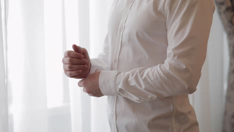 handsome groom man dresses in wedding morning and fixes his buttons on a white shirt sleeve, indoors