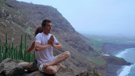 A-man-and-woman-on-a-mountain's-peak-sit-back-to-back-on-a-rock,-meditating-and-doing-yoga,-their-focus-drawn-to-the-ocean-vista