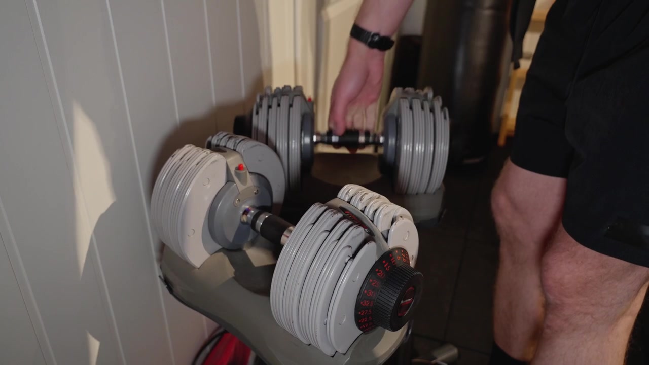 Close up of an adjustable dumbbell rack. A person places both dumbbells back into the rack and walks away