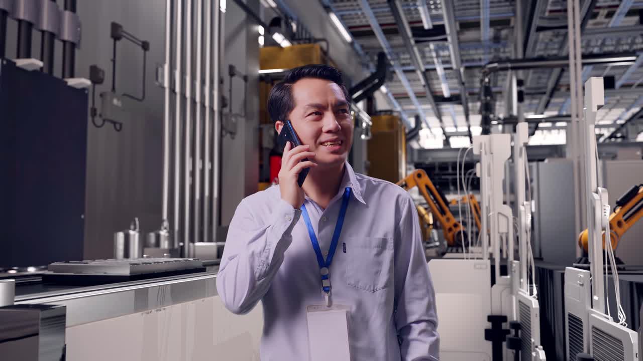 Trabajador profesional asiático de pie, en una línea de producción de baterías automatizada en una fábrica moderna, coge el teléfono y empieza a hablar.