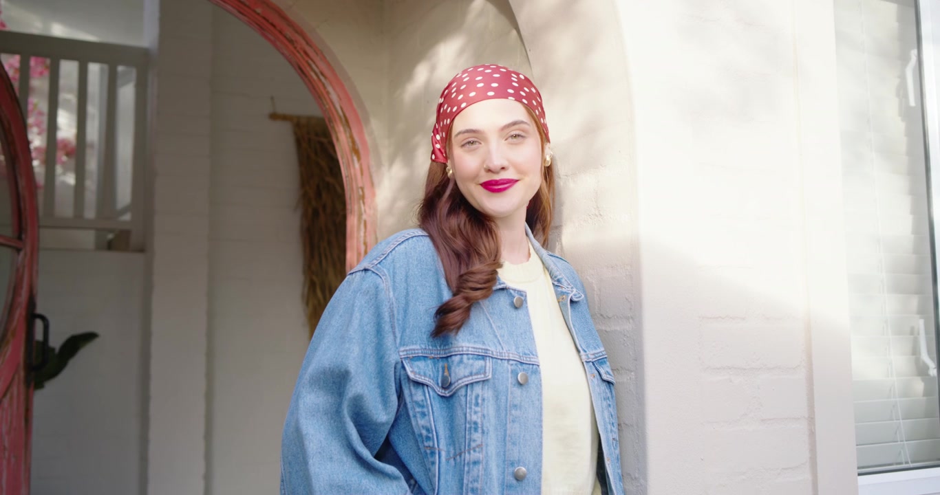 Donna adulta in giacca di jeans appoggiata a una colonna di mattoni su un portico, la luce del sole le illumina il viso mentre sorride e inclina la testa.