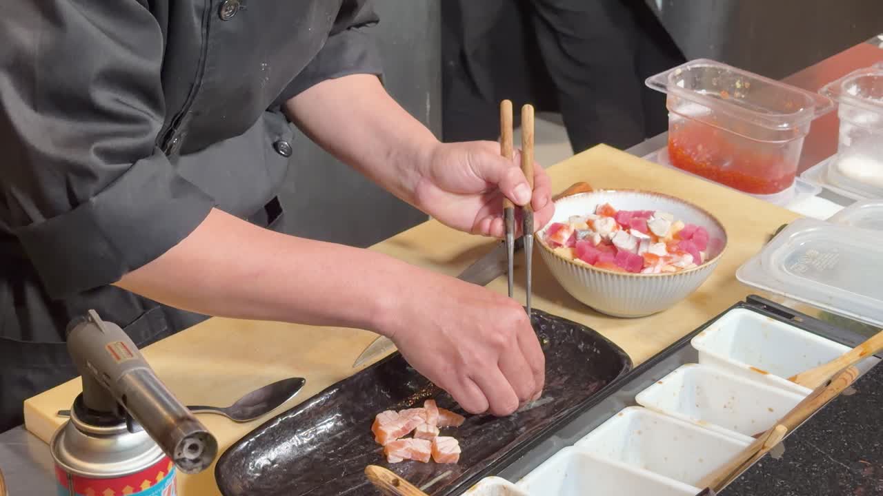 Professioneller Koch richtet Aburi-Lachs-Reis-Bowl mit Stäbchen unter warmer Restaurantküchenbeleuchtung an