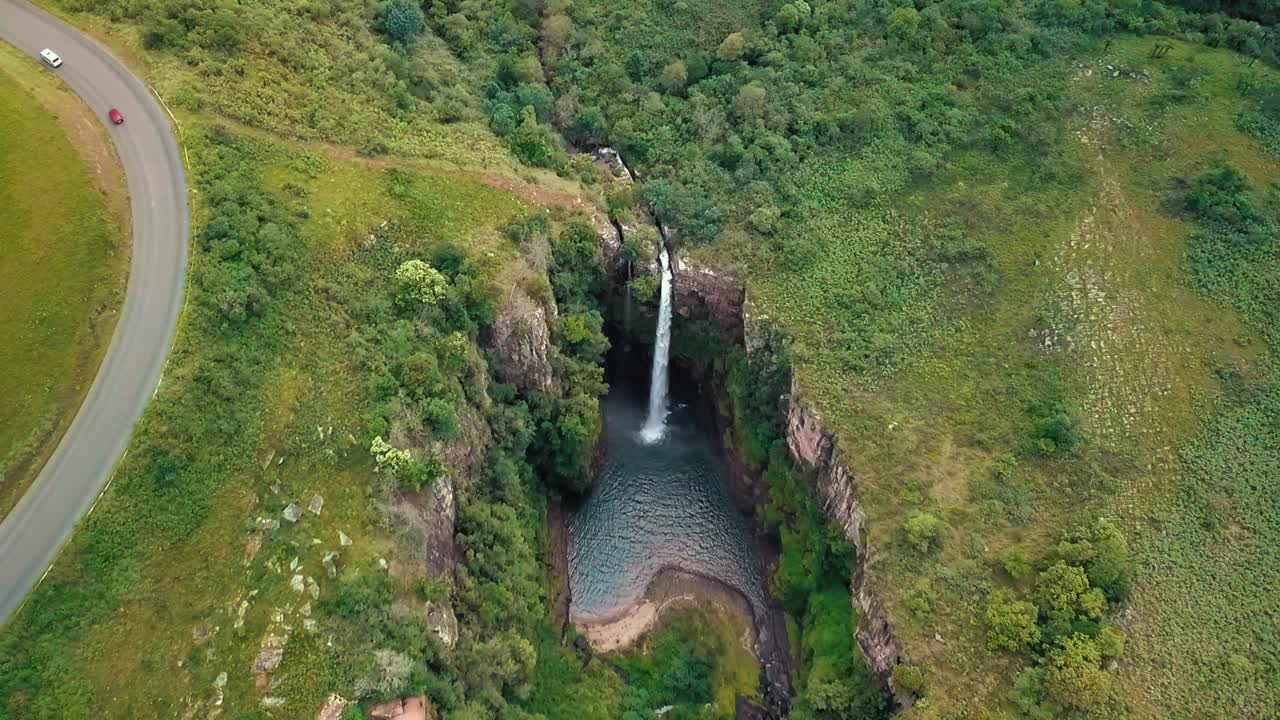 Drone flying over Mac Mac Falls in South Africa showing tall waterfall plunging into rocky pool, surrounded by lush green cliffs, winding road, dense forest, and grassy hills
