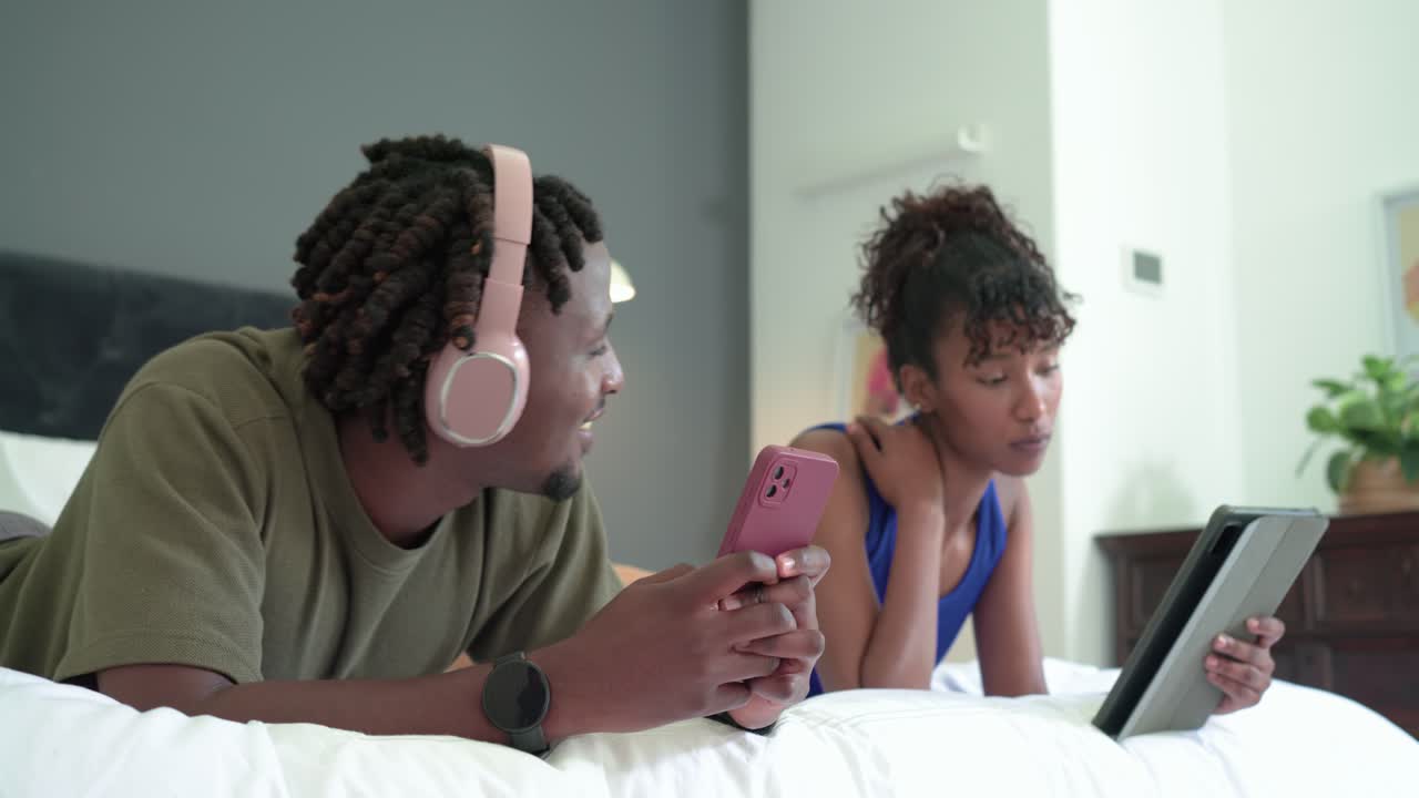 Afroamerikansk par ligger på sengen, hun tapper på tablet og kigger op, han kigger op fra telefonen og smiler