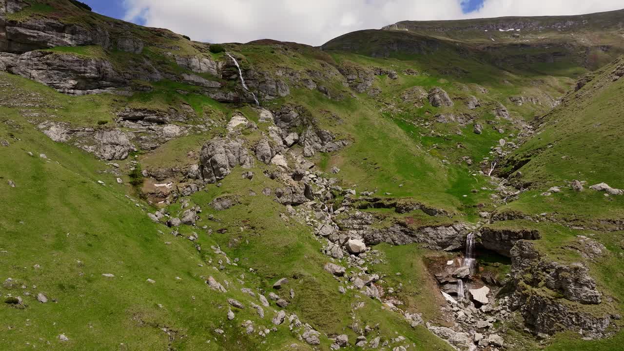 Cascate di montagna a più livelli che scendono da scogliere aspre in una giornata di sole, ripresa con drone in movimento.