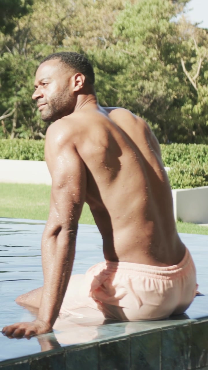 Video verticale: Uomo maturo che riposa, aggiustando la postura sul bordo della piscina dopo una nuotata, con pelle bagnata e costume rosa.