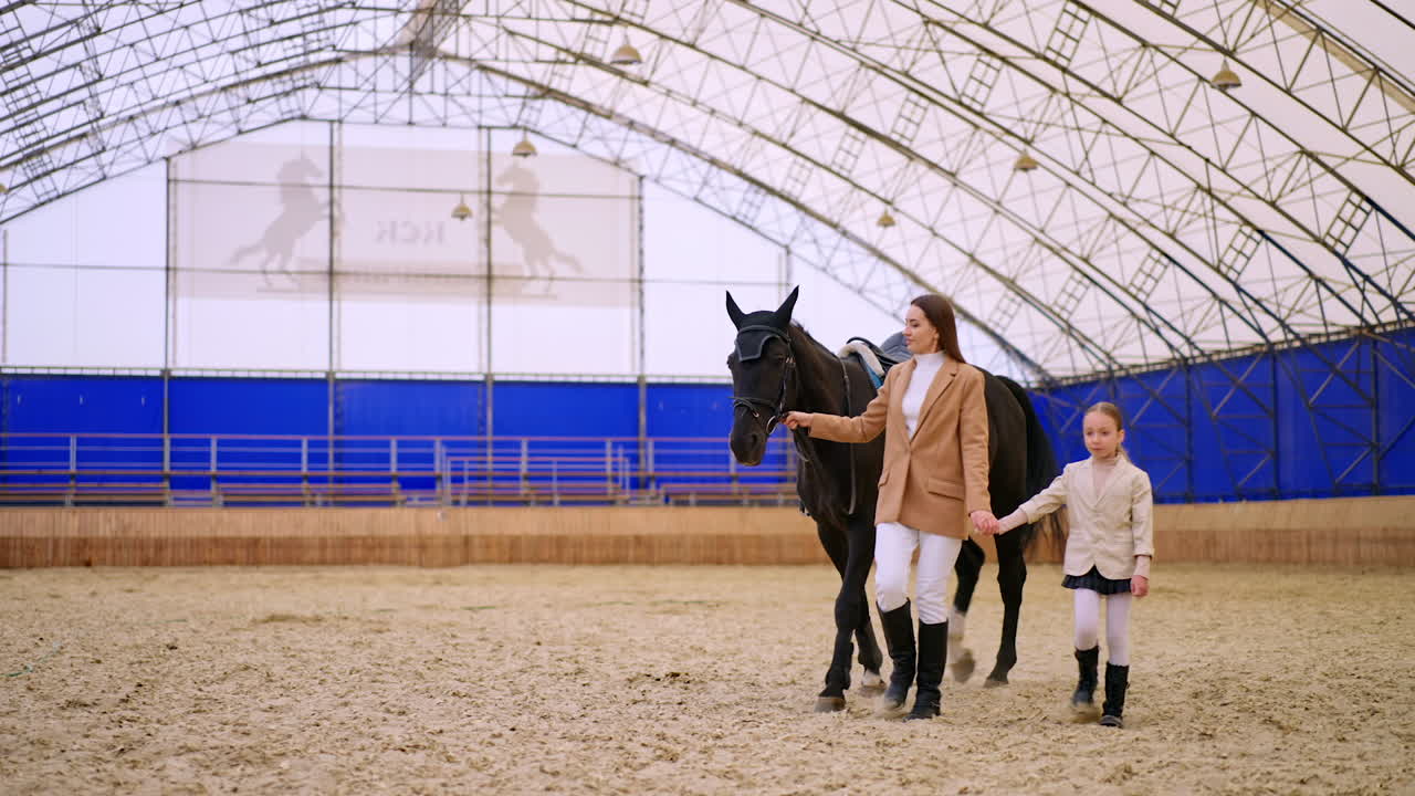 Woman and young girl leading a gorgeous black horse. Mother and daughter spending time at riding hall. Horse riding hobby concept.