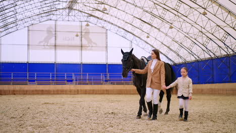 woman and young girl leading a gorgeous black horse. mother and daughter spending time at riding hall. horse riding hobby concept.