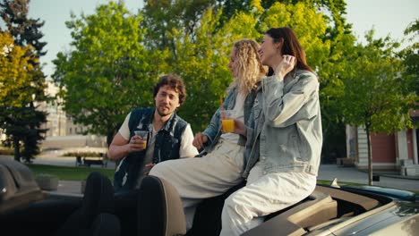 overview of a happy trio two girls and one guy drinking cocktails having fun and chatting on the street near the cabriolet, the girls are sitting on the cabriolet