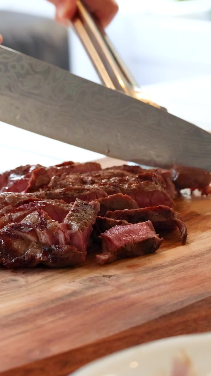 A chef expertly slices a cooked beef steak on a wooden board, showcasing culinary skills in a well-lit kitchen