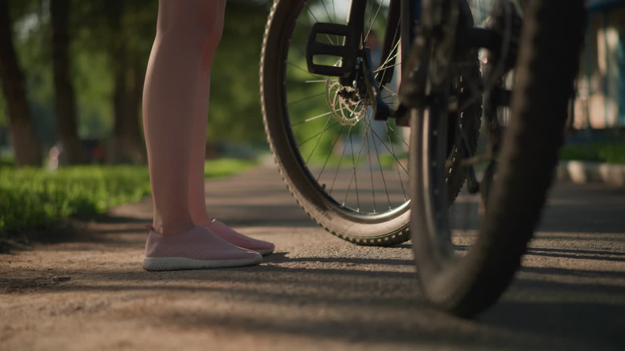 Close-up of lady's legs in pink sneakers kicking a bicycle tire to check air pressure, background showcases lush greenery and trees, casting shadows on the ground while a figure walks by