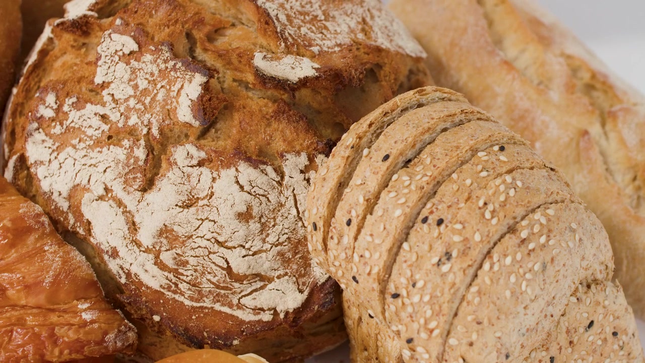 Close-up shot of sourdough loaf, sliced grain bread, and croissants under bright studio lighting.