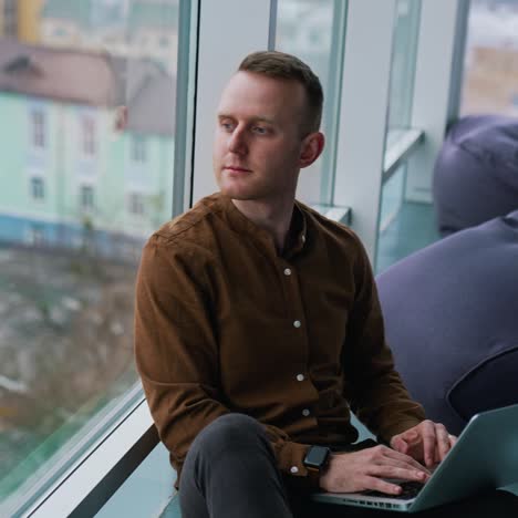Thoughtful man sit on windowsill in office. Businessman with a laptop on laps looking at cityscape through panoramic window daydreaming about future