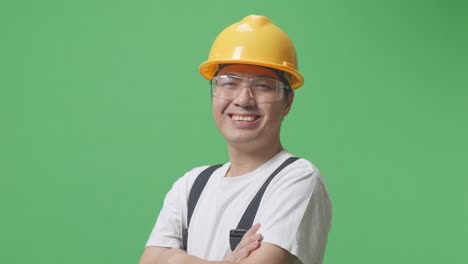 close up of side view of asian man worker wearing goggles and safety helmet smiling and crossing his arms while standing in the green screen background studio