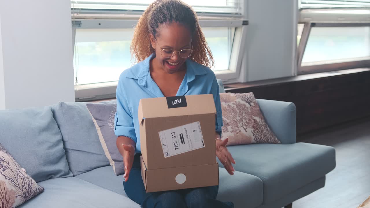Young happy african american woman opens parcel from online store sits on sofa
