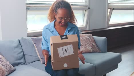 young happy african american woman opens parcel from online store sits on sofa