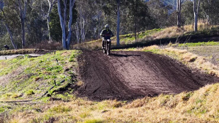 Motociclista da motocross salta su una collina di terra su una pista all'aperto illuminata dal sole, la telecamera segue l'azione in modo costante
