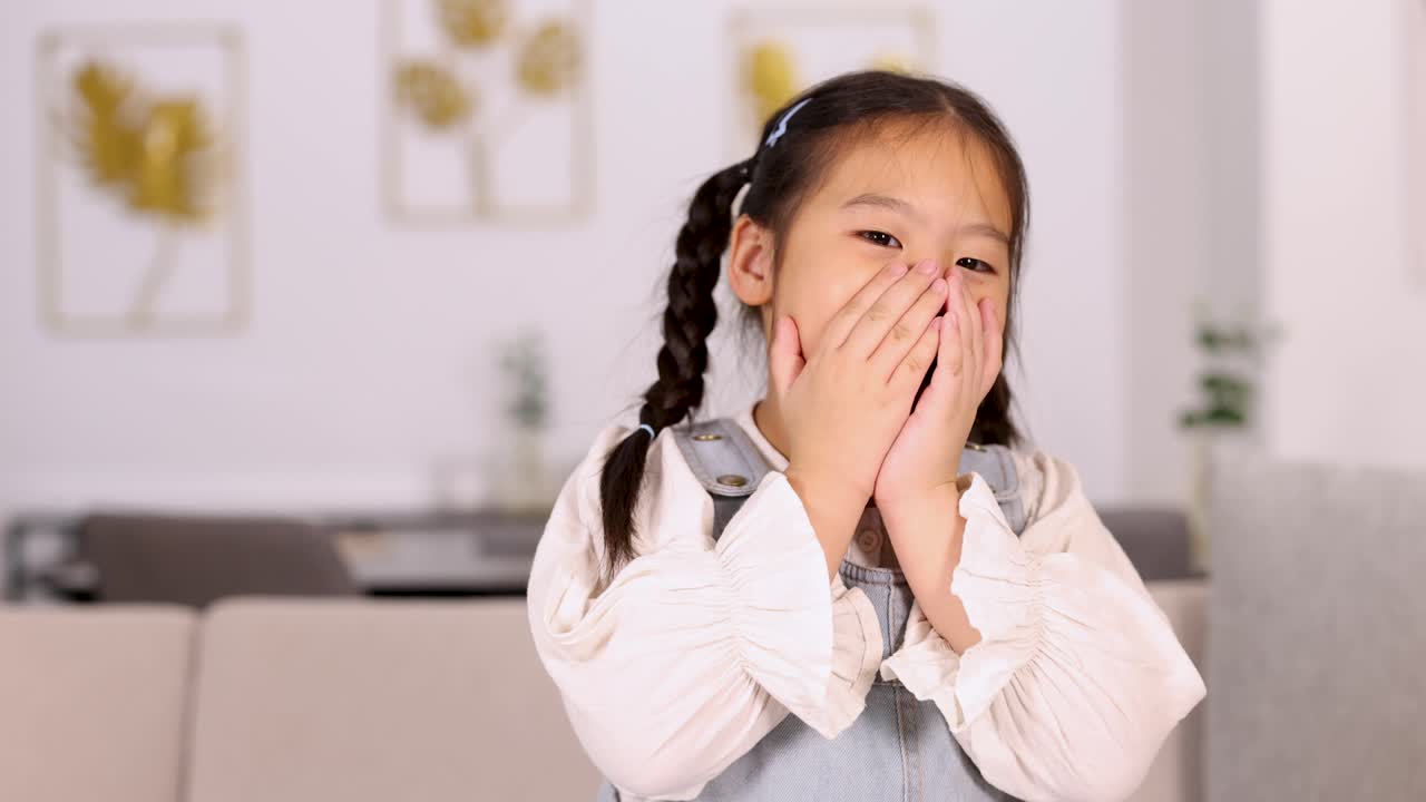 Shy young Asian girl with pigtails smiles in bright, modern living room with natural light