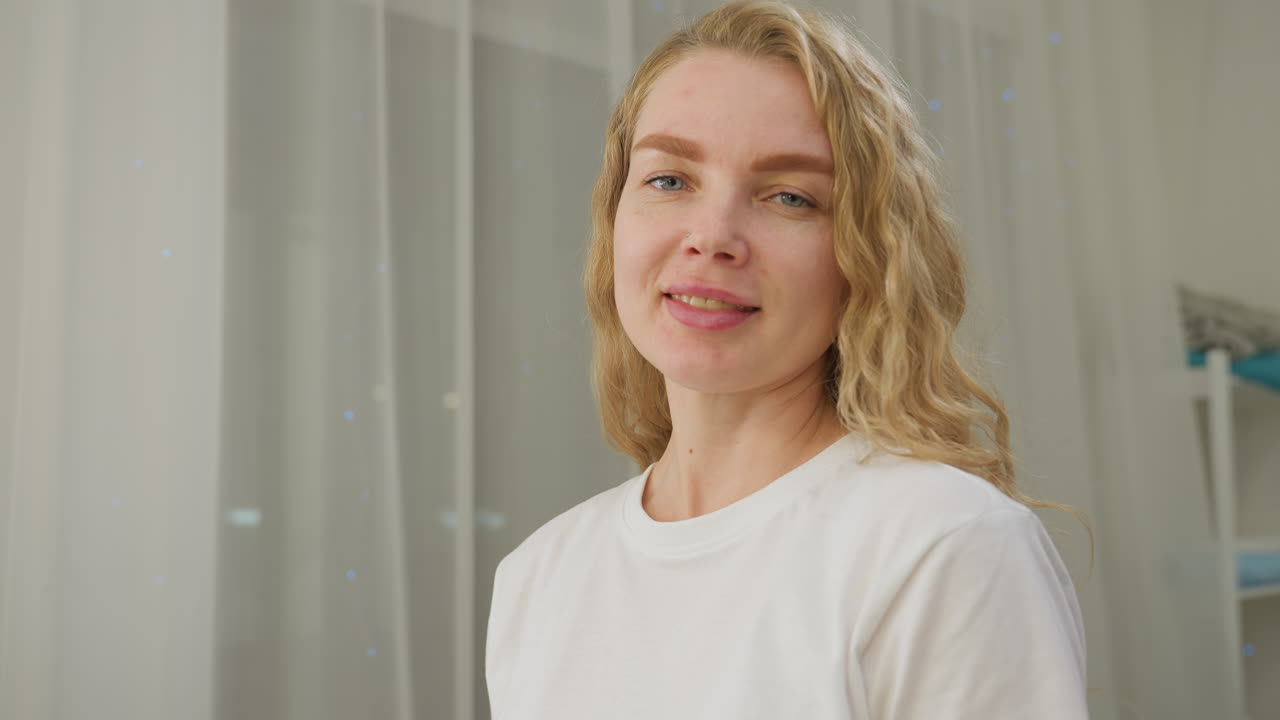 Confident blond woman wearing white shirt smiling gently with subtle nose ring detail, illuminated by soft overhead light and backed by sheer curtains in bright clean setting