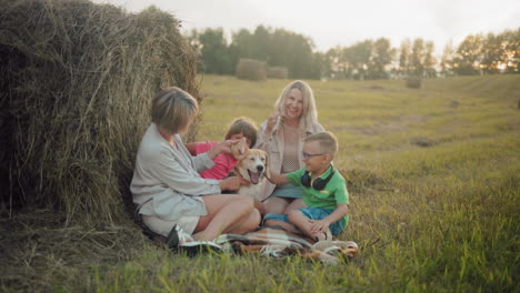 women with children enjoy touching dog in open field, sitting near hay bales during golden hour, relaxed outdoor family time with kids engaging with pet