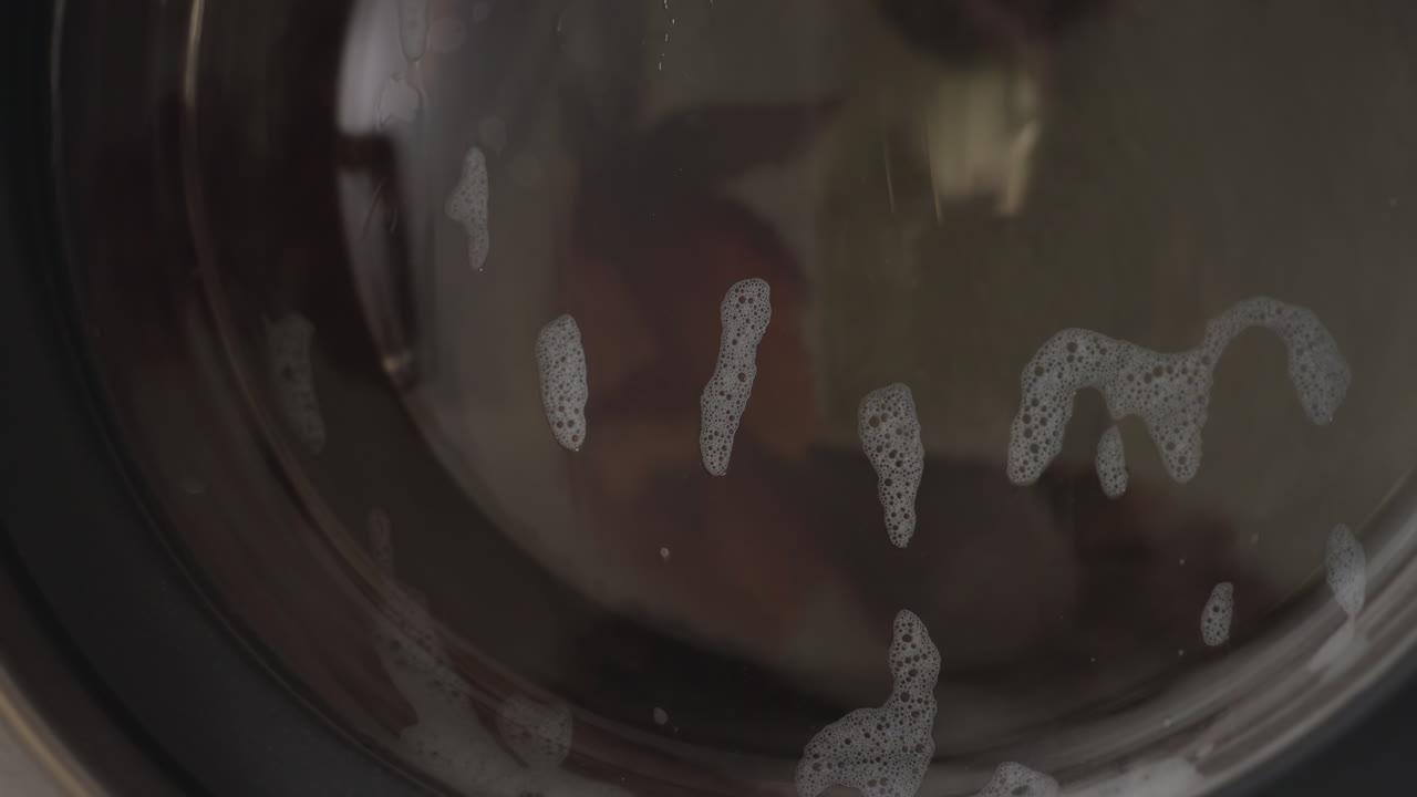 Close up round view of fabric swirling inside washing machine drum, soapy water clinging to glass door, bubbles streaking surface while garments rotate, stainless interior reflecting light