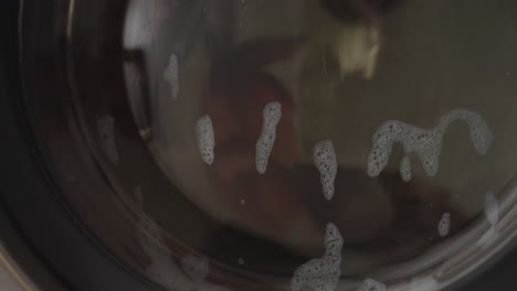 close up round view of fabric swirling inside washing machine drum, soapy water clinging to glass door, bubbles streaking surface while garments rotate, stainless interior reflecting light
