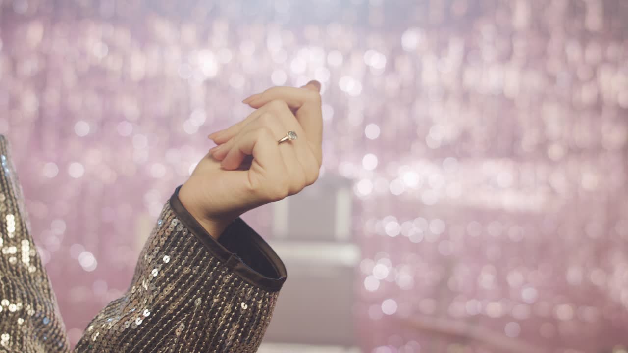 A close-up of a woman's hand, snapping her fingers, wearing a diamond ring and a silver sequin jacket, moving gracefully. The shimmering pink bokeh background creates a glamorous party atmosphere.
