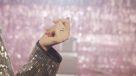 a close-up of a woman's hand, snapping her fingers, wearing a diamond ring and a silver sequin jacket, moving gracefully. the shimmering pink bokeh background creates a glamorous party atmosphere.