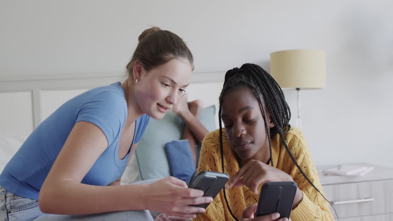 Happy diverse teenage female friends lying on bed talking and using their smartphones