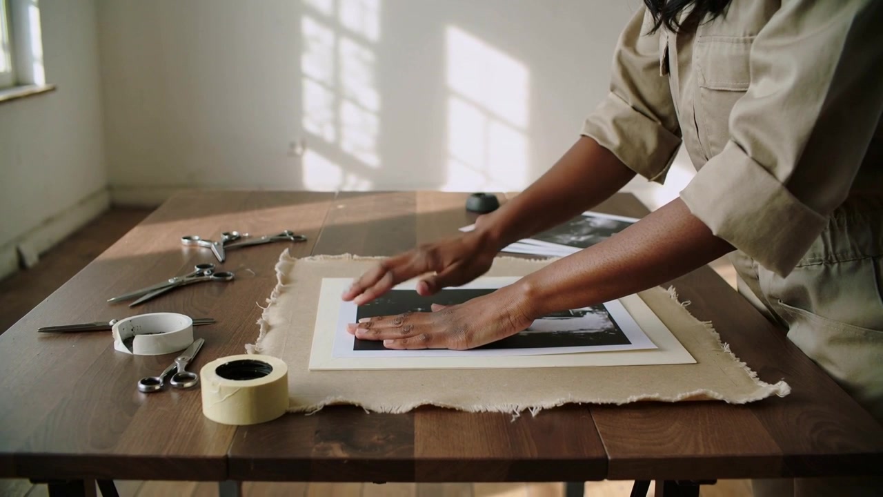 Artista de macacão bege, levantando, posicionando e alisando impressões em um passe-partout pré-cortado para montar com fita adesiva.
