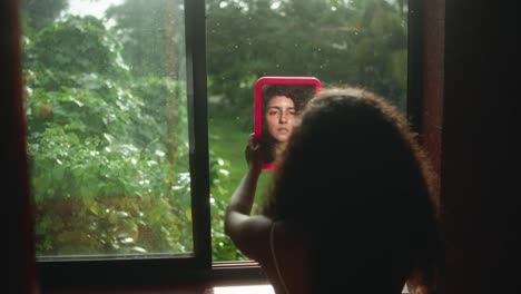 zoom-in on woman’s solemn face in pink mirror, framed by rainy window and lush green jungle outside