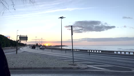 Fantástico-Amanecer-Detrás-Del-Fuerte-De-Carcavelos,-Con-Coches-Circulando-Por-El-Paseo-Marítimo.