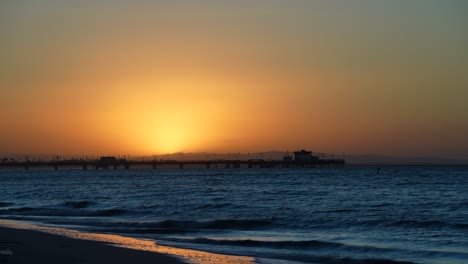 Silhouette-Eines-Piers-Während-Eines-Goldenen-Sonnenaufgangs-Mit-Den-Wellen,-Die-Sanft-über-Einen-Ruhigen-Strand-Spülen---Zeitraffer