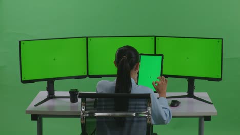 태블릿과 여러 컴퓨터 모니터로 작업하는 아시아 여성의 뒷면은 녹색 스크린 스튜디오에서 녹색 화면 모형을 가지고 있습니다.