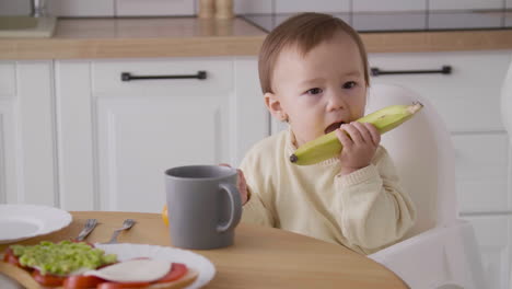 Linda-Niña-Sentada-En-Una-Silla-Alta-Al-Lado-De-La-Mesa-De-La-Cocina-Sosteniendo-Y-Mordiendo-Un-Plátano