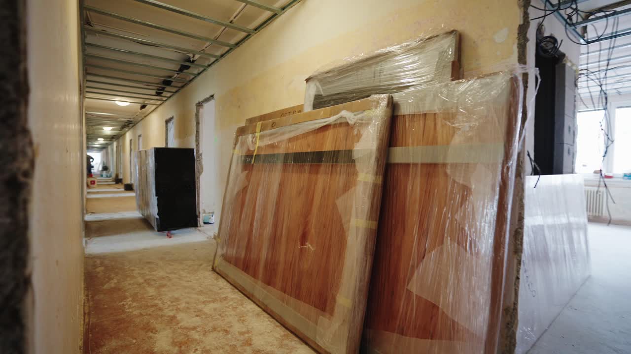 New wooden doors wrapped in protective plastic leaning against a wall in an unfinished hallway during apartment renovation