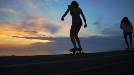 As-the-sun-sets,-two-friends-in-shorts-ride-skateboards-along-a-road,-capturing-the-mountains-and-a-stunning-sky-in-the-background,-all-in-slow-motion