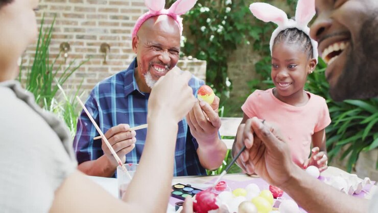 Happy african american parents, daughter and grandfather painting easter eggs in garden, slow motion
