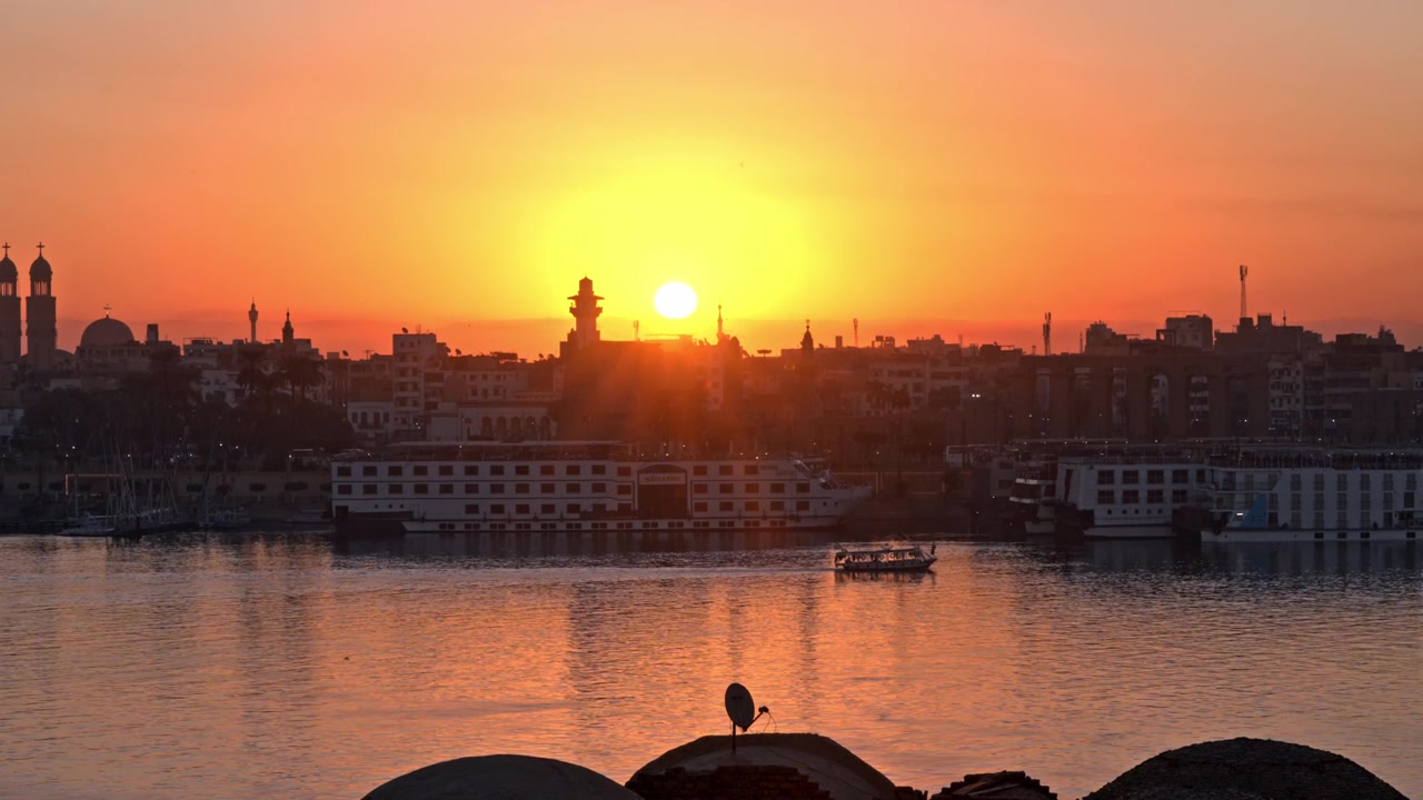 Intenso bagliore dorato del sole che tramonta direttamente sullo skyline di Luxor, proiettando riflessi brillanti sul fiume Nilo dove navi da crociera e imbarcazioni locali riposano lungo la Sponda Orientale