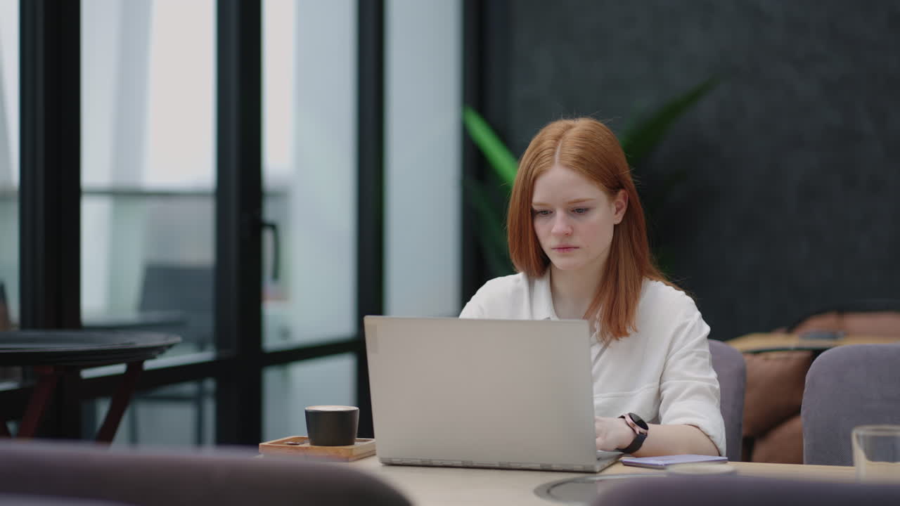 A young red-haired woman is working on a laptop. Remote work. Home office. Office work. A woman in a suit in the office is typing on a laptop