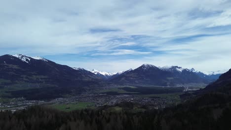 Drone-flies-above-pine-forest-towards-town-surrounded-by-snow-covered-Mountains-in-Nenzing,-Voralberg,-Austria