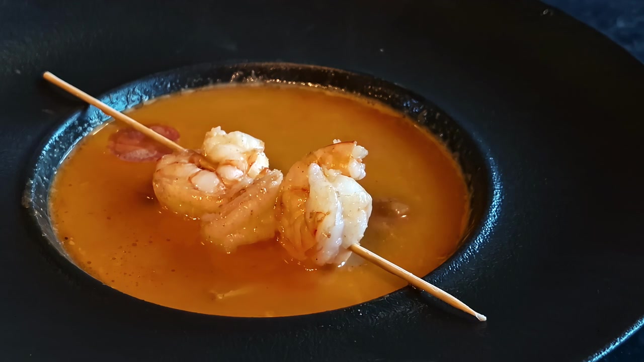 En fyldig, orange skaldyrssuppe serveret med et træspyd med to grillede rejer.