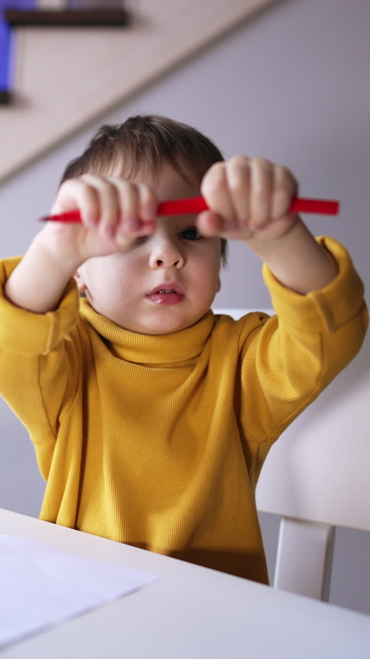 Small toddler in yellow sweater sitting at desk. Kid holds a red pen and waving it. Child drawing at home. Vertical video