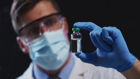 médico caucásico con máscara protectora y guantes sosteniendo el vial de vacuna