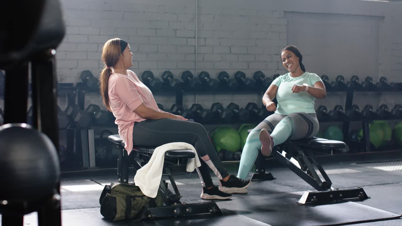 Diverse female pals in gym mint woman raising hand sparking high fives laughing checking smartwatch