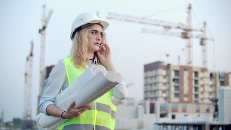 Sprechende-Frau-Mit-Helm-Am-Telefon-Vor-Dem-Hintergrund-Einer-Baustelle-Mit-Kränen,-Die-Zeichnungen-In-Der-Hand-Halten.-Ingenieurin-Auf-Der-Baustelle